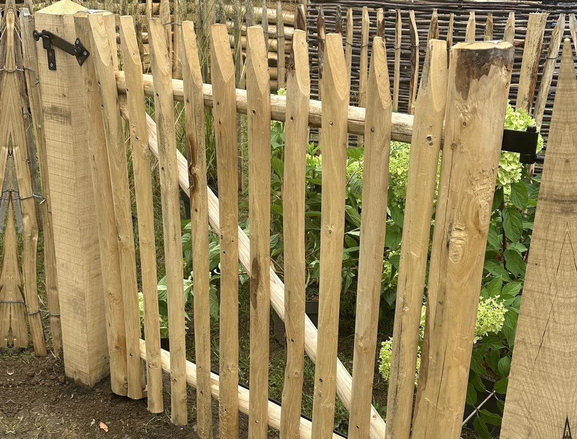 Gemonteerd kastanjehouten tuinpoortje met gepunte latten en contrasterend zwart beslag in een landelijke tuin.