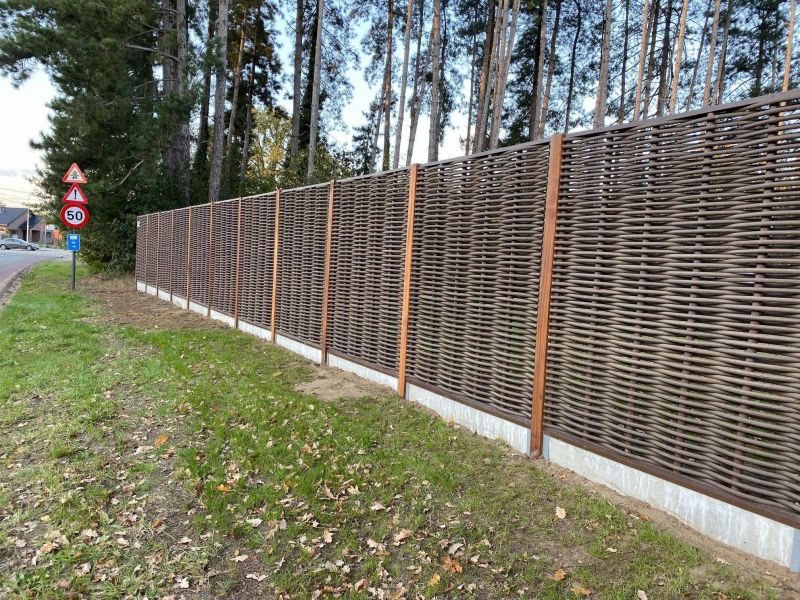 Forest Avenue gevlochten tuinscherm van kunststof fiber in een natuurlijke bruine kleur, geplaatst op een betonplint langs een bosrand.